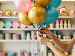 Handen die in een feestartikelenwinkel een ballonnenboeket in roze, goud en hemelsblauw samenstellen, versierd met krullend lint.