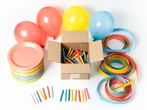 Overhead flat lay of colorful birthday party supplies including paper plates, streamers, balloons, and candles in coral, yellow, and blue around a wholesale box.