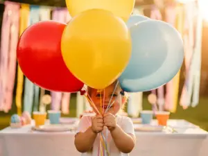 Smiling child holding red, yellow, and blue latex balloons at a sunny outdoor birthday party with pastel streamers in the background.