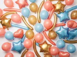 Overhead flat lay of colorful latex, foil star, heart-shaped, and twisting balloons in coral pink, gold, and sky blue on a white surface.
