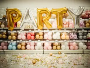 Gouden en zilveren letter die samen een naam vormen, zweven boven kleurrijke ballonnenpakketten in een feestartikelenwinkel, terwijl er feestelijke confetti op de vloer ligt.