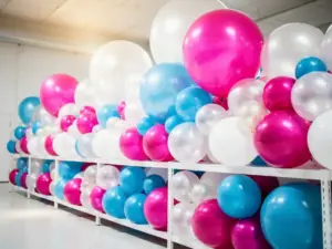 Colorful latex balloons in pearl white, magenta, and sky blue arranged on a warehouse shelf, ranging from 5-inch rounds to 36-inch statement sizes.