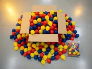 Bulk wholesale latex balloons in red, yellow, and blue spilling from an open cardboard box beside a small retail bag for scale.