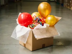 Colorful party balloons and streamers packed in a kraft cardboard shipping box with bubble wrap and tissue paper padding.