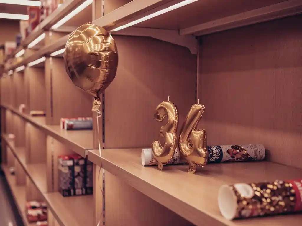 Una estantería casi vacía en una tienda de artículos para fiestas, con un único globo de papel metalizado, Velas con números gigantes Velas con números y cañones de confeti, todo ello bañado por una cálida luz ámbar.