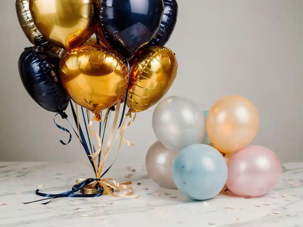 Metallic gold and navy foil balloon bouquet with satin ribbons beside simple pastel latex balloons on white marble, soft confetti scattered around both.