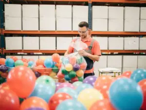 Warehouse worker inspecting colorful latex balloons in bulk, with stacked wholesale boxes in a bright modern distribution facility.