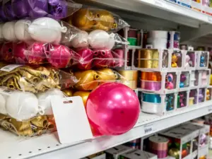 Artículos para fiestas al por mayor, como globos de látex apilados, rollos de cinta y adornos en cajas, sobre una estantería blanca de almacén con una etiqueta de precio escrita a mano.