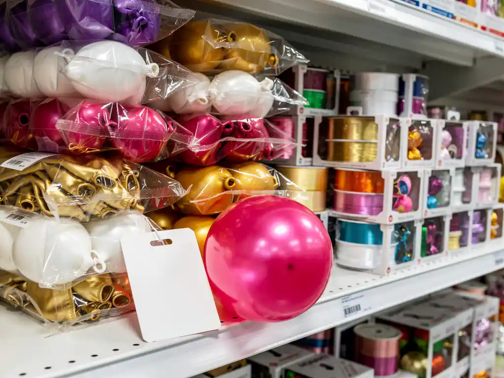 Wholesale party supplies including stacked latex balloons, ribbon rolls, and boxed decorations on a white warehouse shelf with a handwritten price tag.